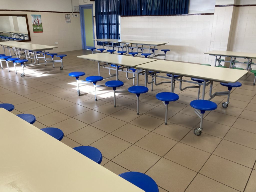 Comedor escolar vacío, con mesas alargadas y taburetes azules abatibles