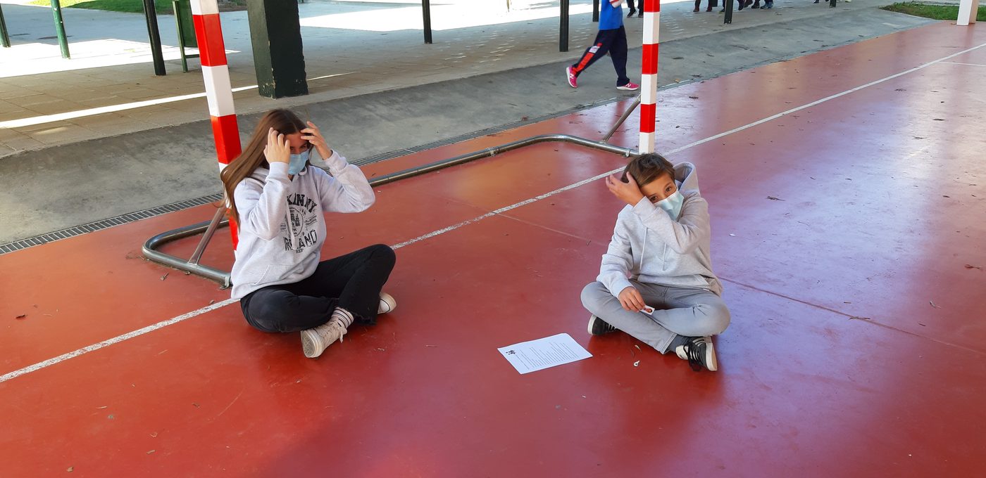 Dos alumnas con mascarilla sentadas en el suelo de la pista, separadas, con una hoja entre ellas