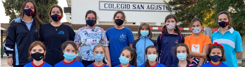 Grupo de alumnas y monitoras posando con mascarilla ante el rótulo del colegio