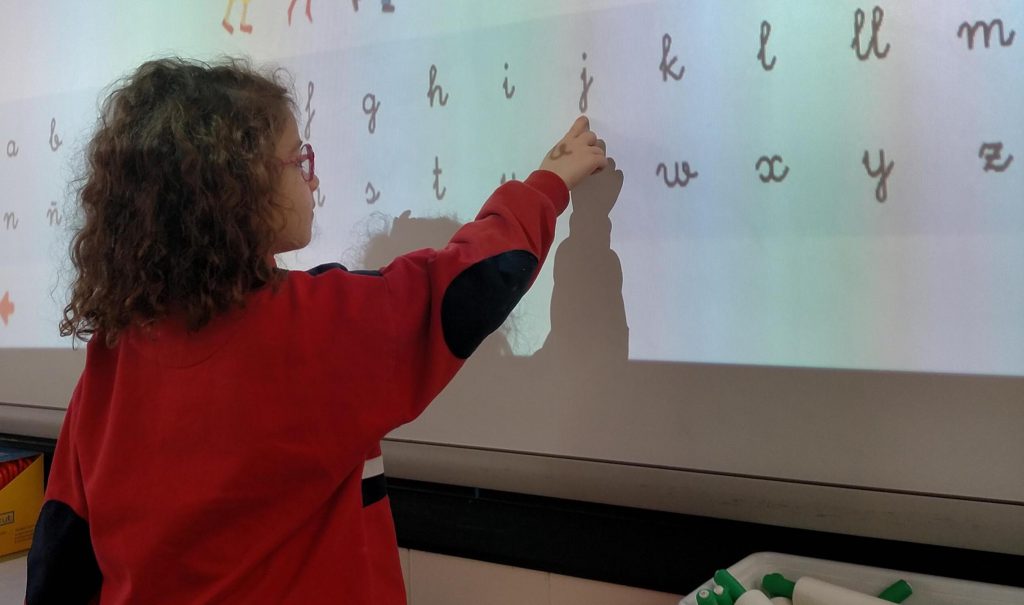 Niña señalando letras del abecedario en cursiva proyectadas en la pizarra