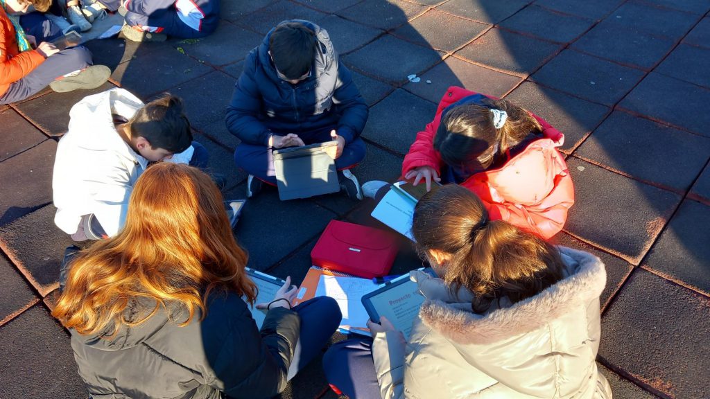 Alumnos sentados en el suelo del patio, en corro, trabajando con tabletas