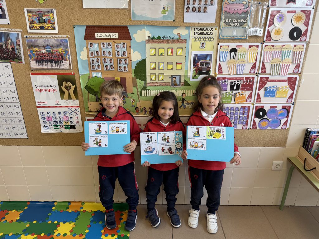 Tres niños de Infantil mostrando sus trabajos delante del panel de la clase