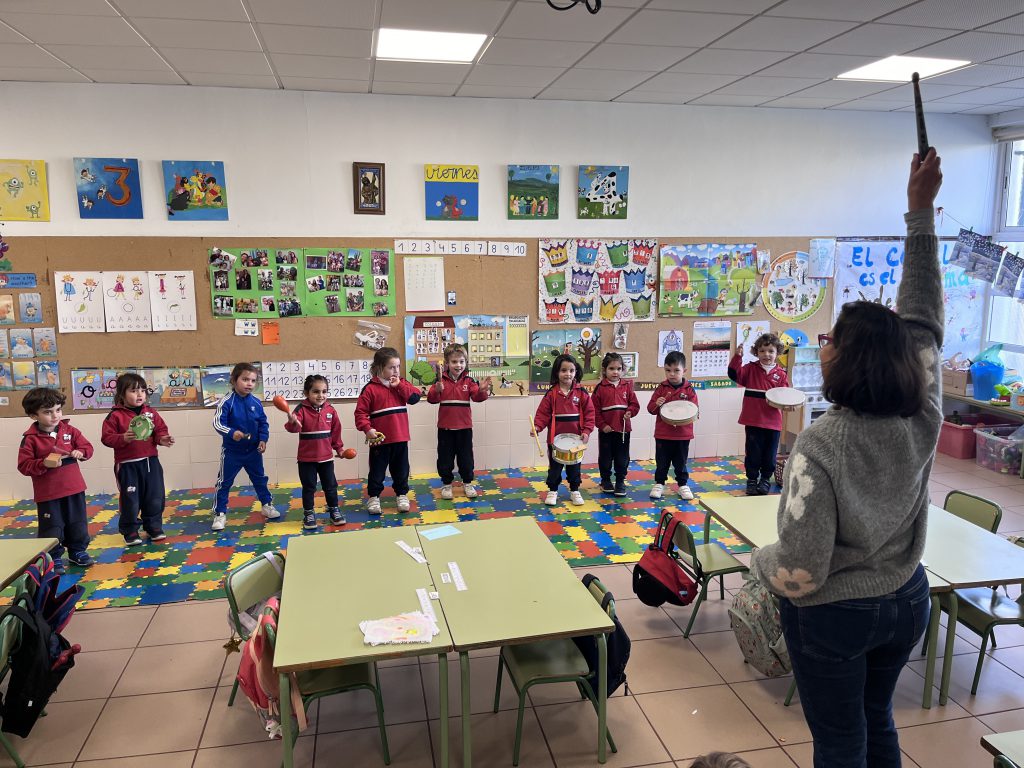 Aula de Infantil con los niños de pie sobre la alfombra y la maestra dirigiendo