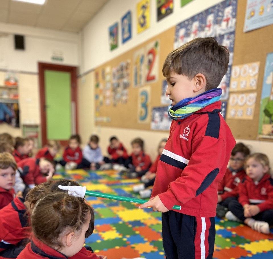 Niños de Educación Infantil sentados en la alfombra de colores del aula, atentos a un compañero que está de pie