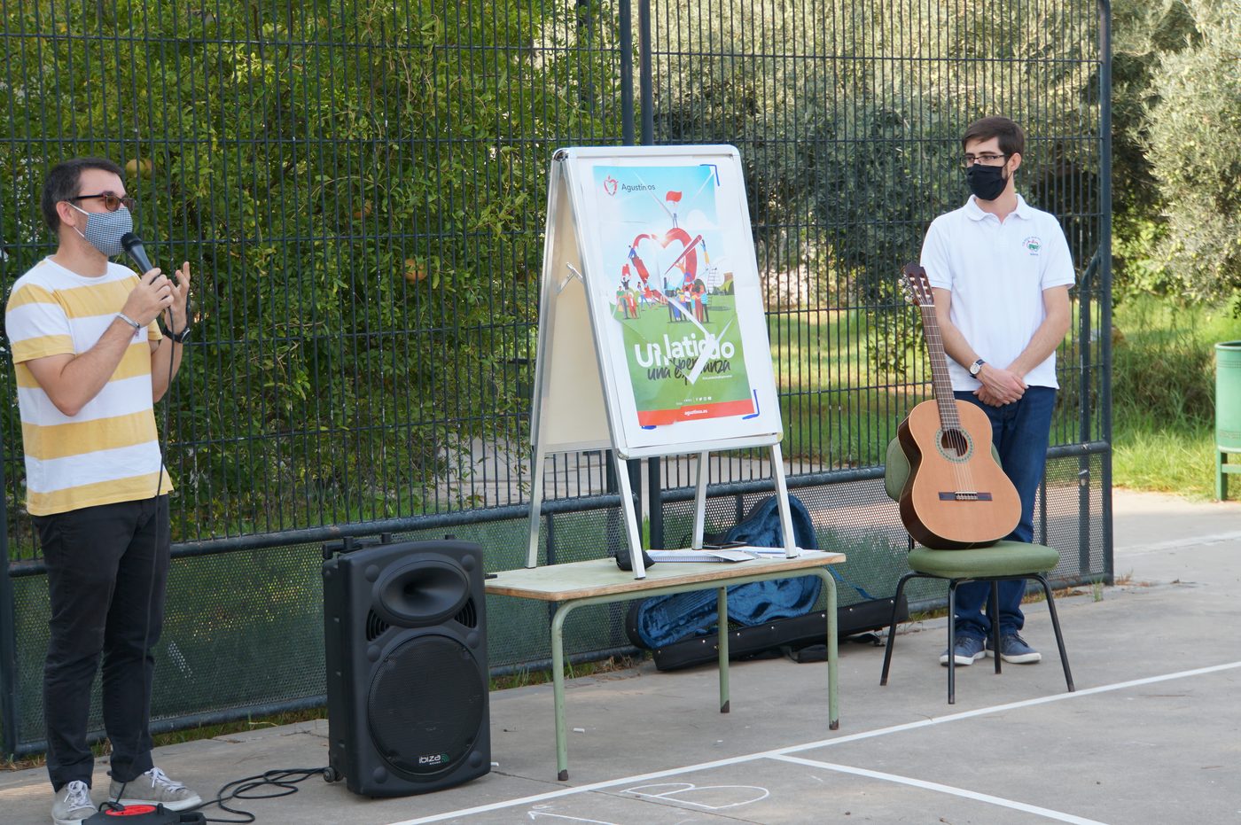 Dos profesores con mascarilla, uno con micrófono y otro junto a una guitarra, ante un cartel con el lema «Un latido, una esperanza»
