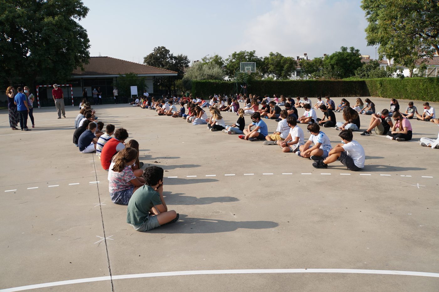 Cientos de alumnos sentados en el suelo del patio, separados unos de otros, durante un acto al aire libre