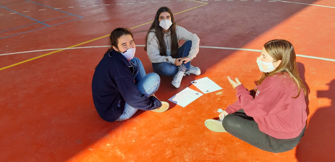 Tres alumnas con mascarilla sentadas en el suelo de la pista, en triángulo, trabajando sobre unos papeles