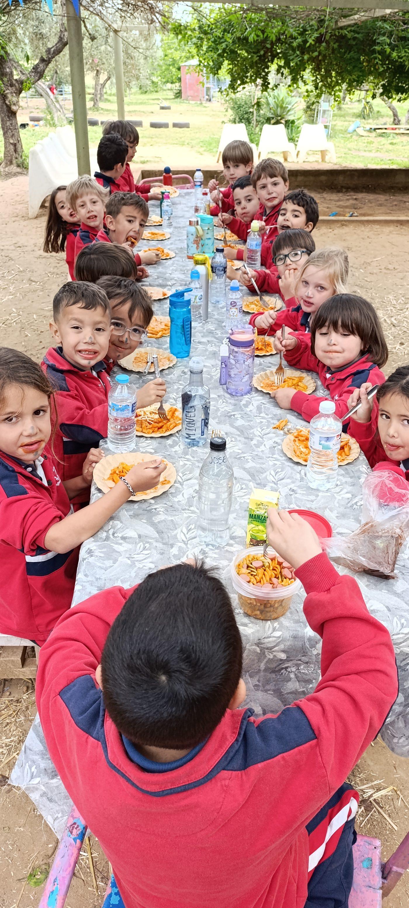 Niños y niñas de Infantil desayunando en una mesa larga bajo los árboles