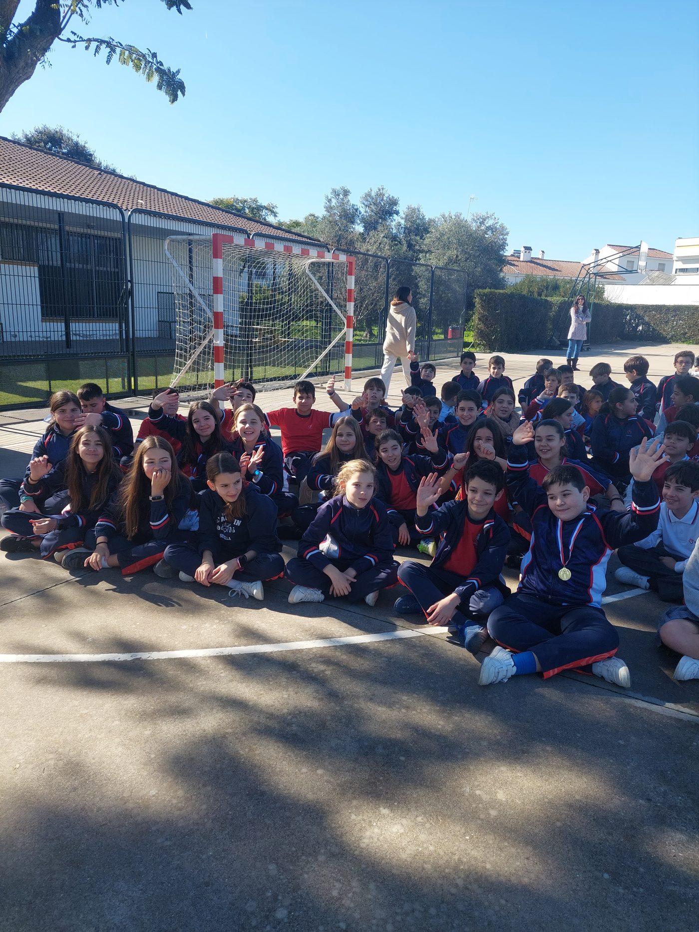 Grupo numeroso de alumnos sentados en el suelo del patio, al sol, saludando a la cámara